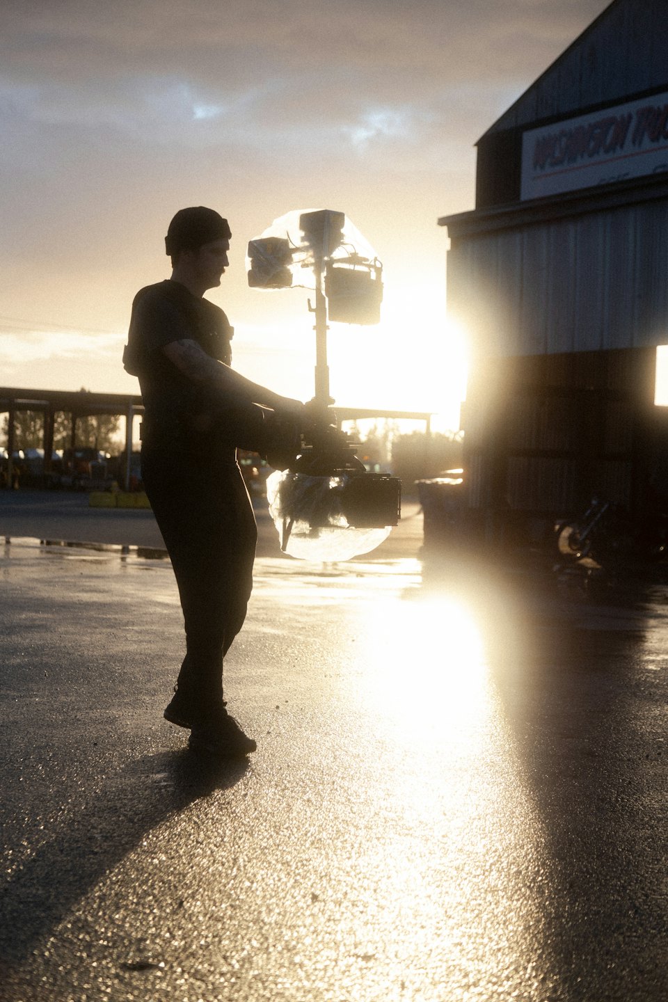 Film crew member silhouetted operating camera gimbal on wet asphalt.